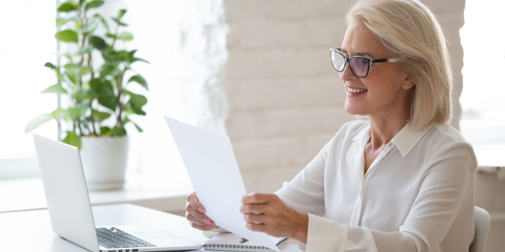 Smiling middle-aged woman in glasses sit at office desk work at laptop